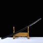 Diagonal view of a Japanese cosplay katana displayed on a carved wooden stand, photographed against a solid black background with a light-colored floor. The glossy black lacquered shirasaya scabbard is fully visible, decorated with silver cherry blossom motifs, while the blade is not visible in this shot. The sheath is present with a minimalist shirasaya design intended for decorative and cosplay display.
