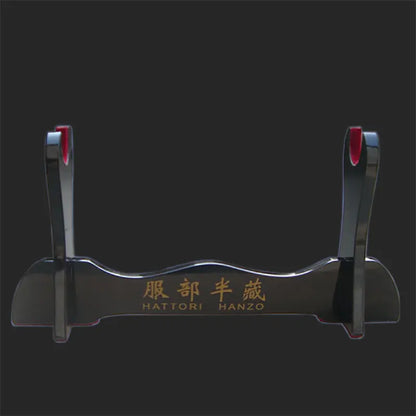 Présentoir pour katana en bois laqué noir, modèle Hattori Hanzo, photographié sur un fond noir uni, montrant le support brillant avec inscription dorée et protections rouges, utilisé comme support de sabre japonais, kake décoratif pour exposer katana, wakizashi ou shirasaya.