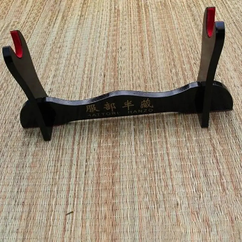 Présentoir pour katana en bois noir laqué, modèle Hattori Hanzo, assemblé et posé sur un tapis en fibres naturelles, montrant les montants avec encoches rouges et la base incurvée avec inscription dorée, utilisé comme porte-katana ou kake japonais pour exposer un sabre traditionnel.