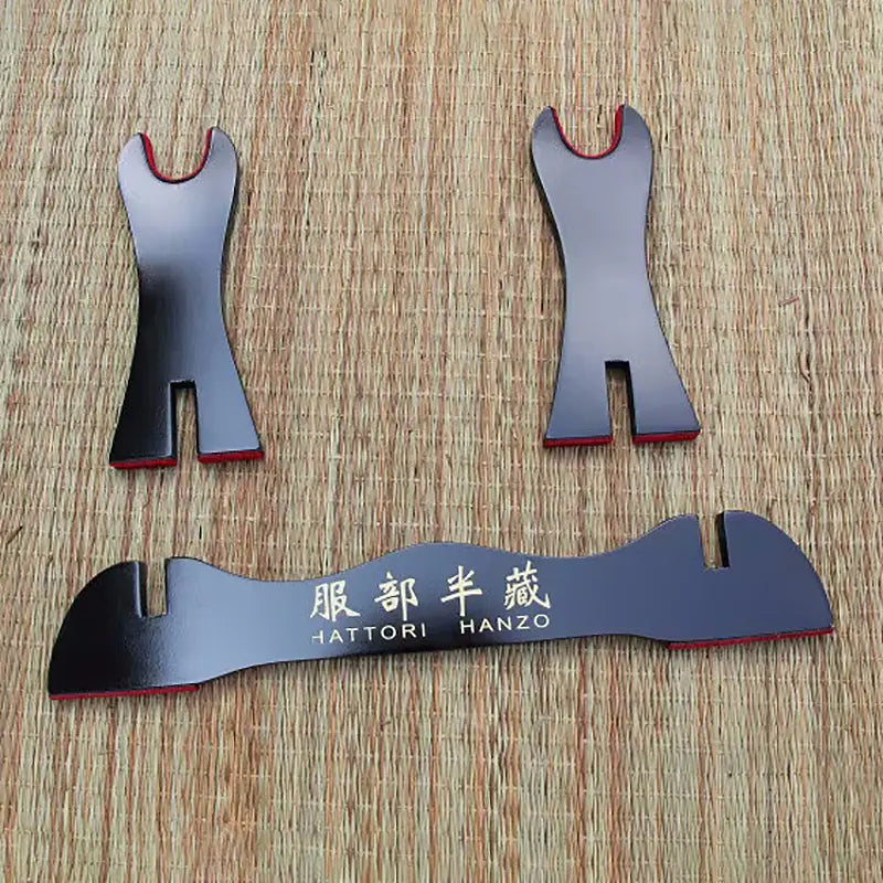 Ensemble de pièces détachées d’un présentoir pour katana en bois laqué noir, comprenant la barre frontale avec inscription dorée “Hattori Hanzo” et les deux montants latéraux, posés sur un tapis en fibres naturelles, montrant les éléments du kake japonais avant assemblage pour exposer un sabre traditionnel.