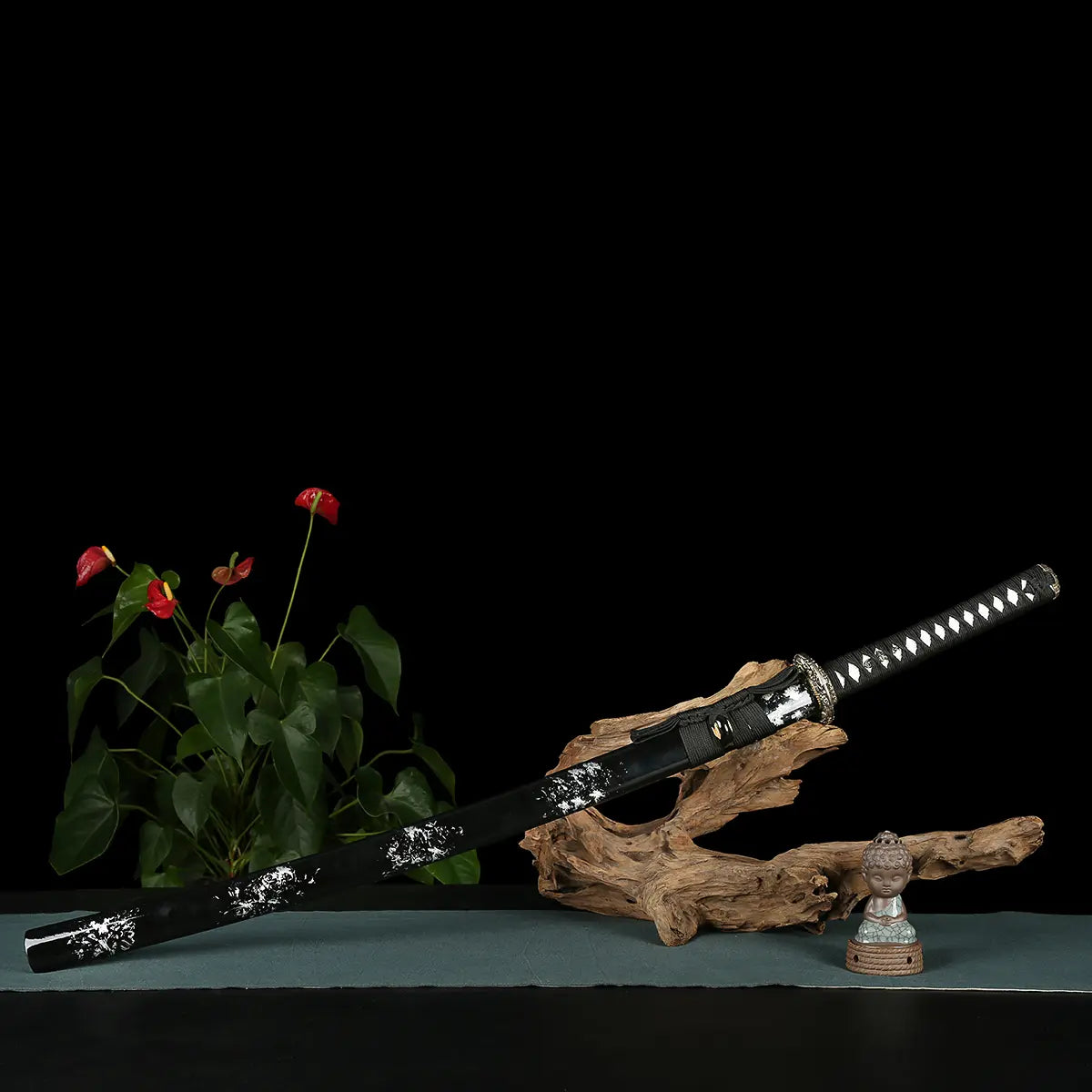 Japanese handmade katana Renge no Ken displayed on a zen-style setup against a deep black background, black glossy saya with white lacquer splash patterns resting on natural wooden driftwood, katana fully sheathed, black tsuka wrapped with black cord over white ray skin, lotus-pattern tsuba visible, green plant and small Buddha statue placed beside the sword for a calm traditional atmosphere
