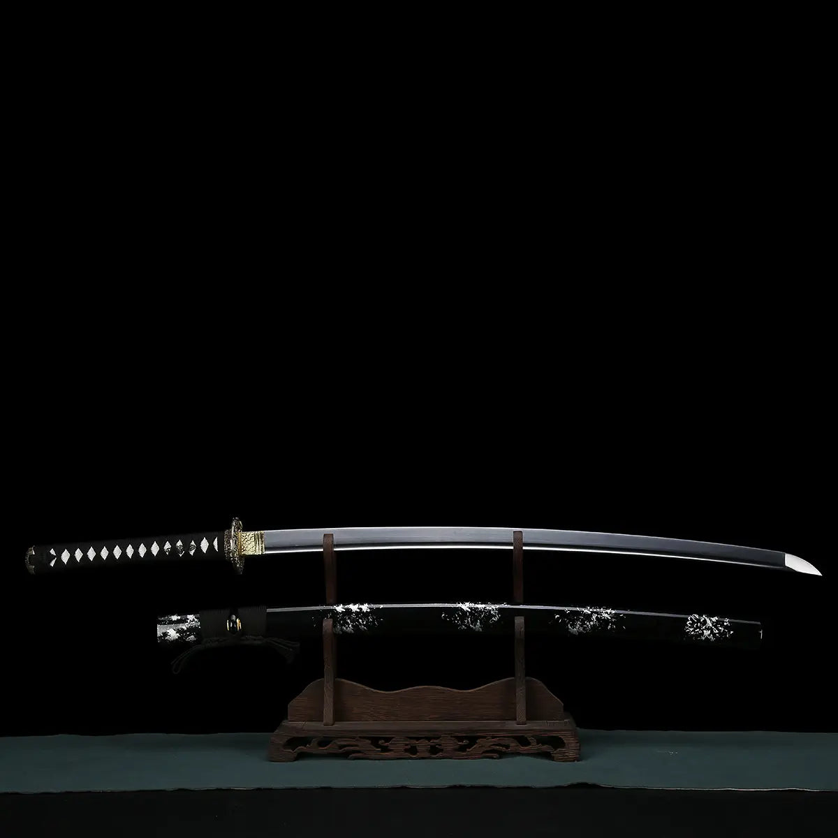 Japanese handmade katana Renge no Ken displayed fully unsheathed on a wooden stand against a deep black background, polished carbon steel blade with visible bohi groove, sharp kissaki, black tsuka wrapped with black cord over white ray skin, gold-toned habaki and dark metal lotus-pattern tsuba, traditional Japanese sword presented horizontally
