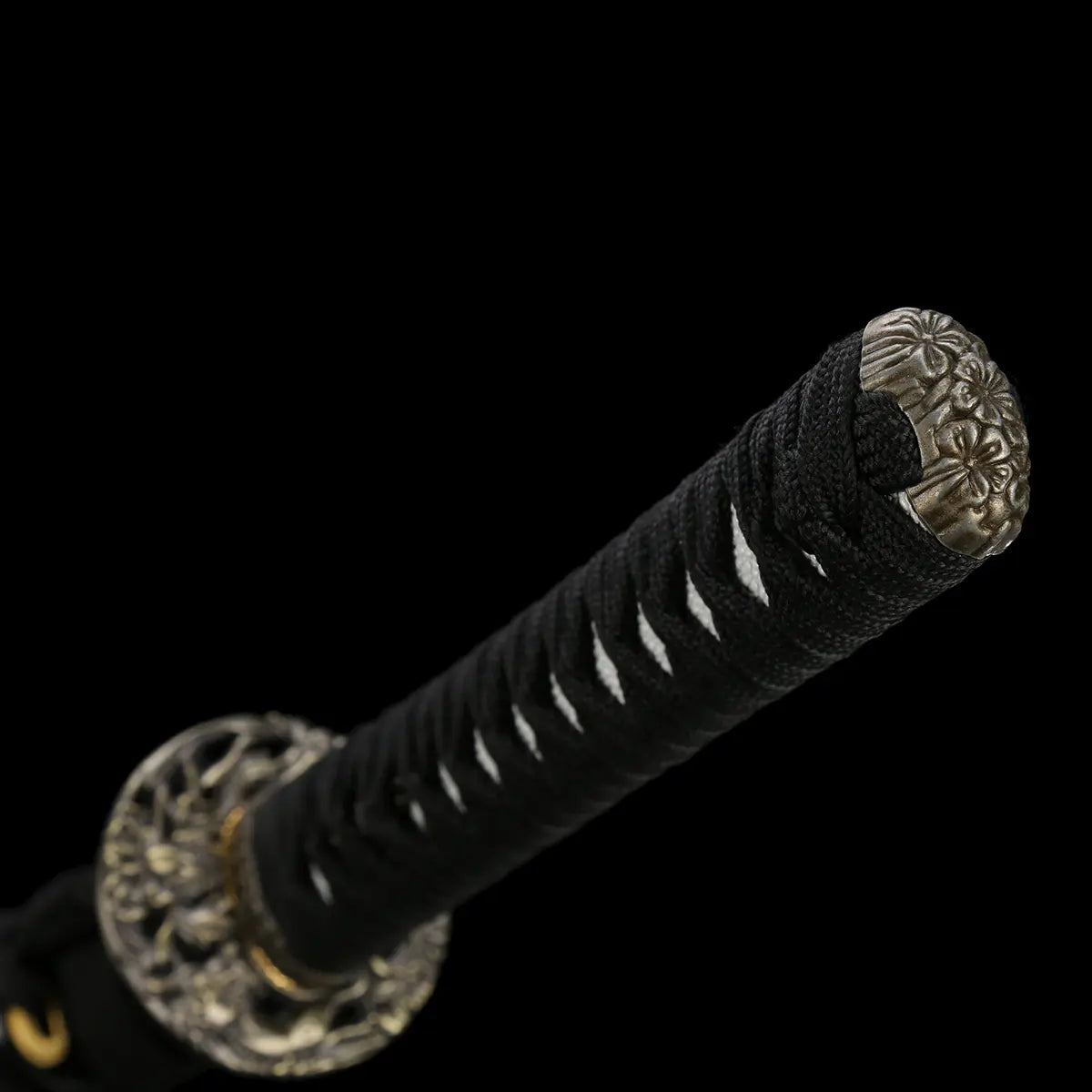 Close-up of the handle of the Japanese handmade katana Renge no Ken on a deep black background, black tsuka wrapped with black cotton cord over white ray skin, lotus-pattern kashira in dark metal, matching lotus motif tsuba partially visible, traditional Japanese sword handle details shown without scabbard
