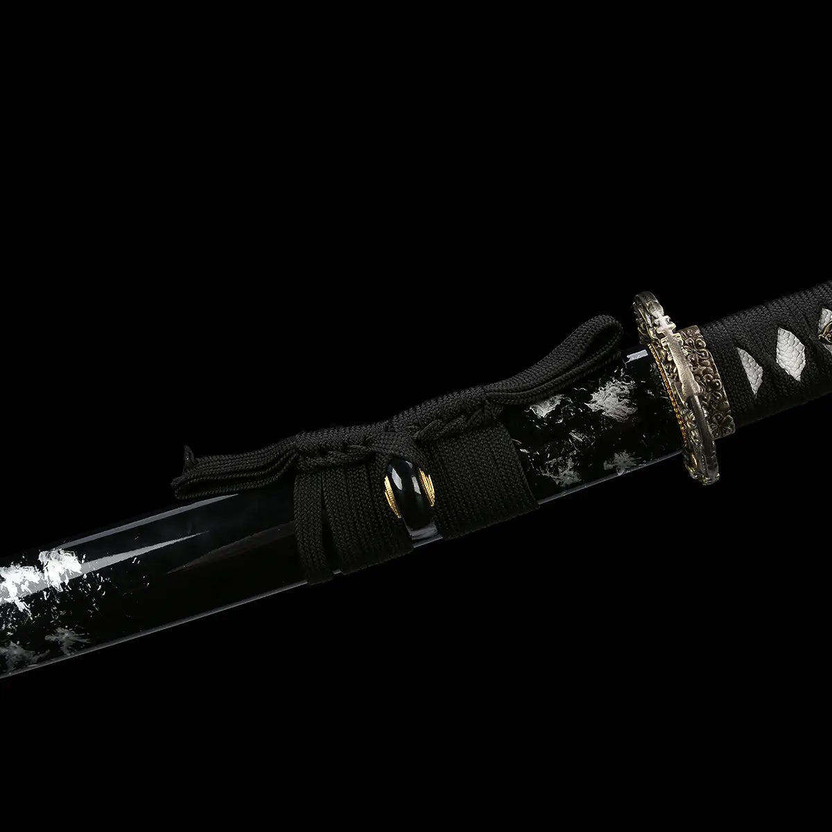 Close-up of the Japanese handmade katana Renge no Ken on a deep black background, black glossy saya with white lacquer splash patterns, katana partially sheathed, black sageo cord tied around the scabbard, dark metal lotus-pattern tsuba and gold-accented habaki visible, traditional Japanese sword fittings shown in detail
