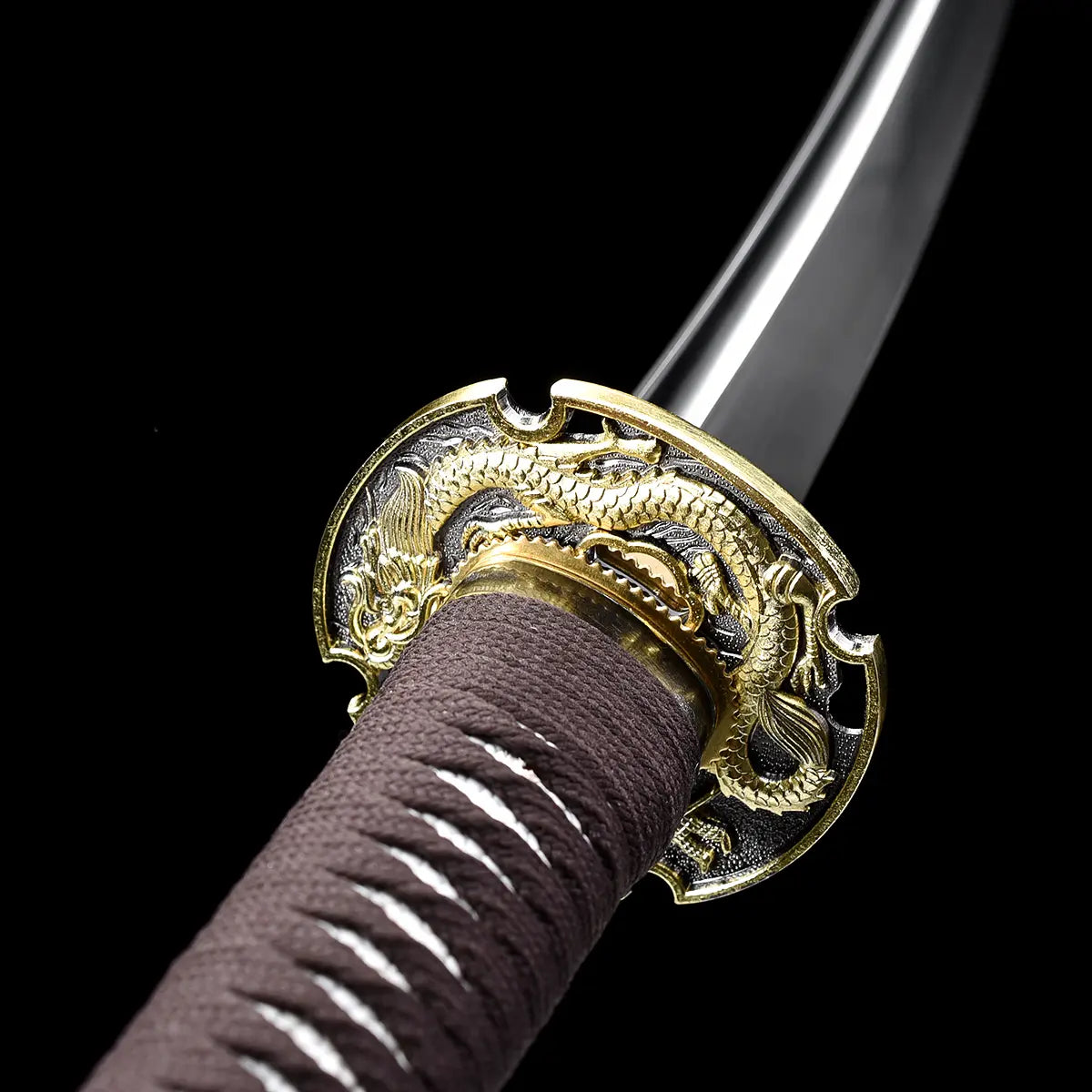 Close-up of the Ryusei Japanese katana focusing on the dragon-themed tsuba and the base of the blade, photographed against a solid black background. The gold-colored dragon guard shows detailed engraved relief, paired with a matching gold habaki. The blade in 1060 carbon steel appears polished and slightly curved, while the handle is wrapped in dark brown cord over white ray skin panels, highlighting the traditional materials and precise fittings of this Japanese katana.
