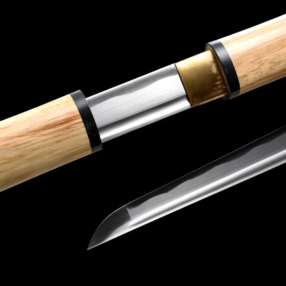 Gros plan du tantō shirasaya San-Ken : habaki en laiton, saya/poignée en bois clair avec bague noire, lame polissage miroir avec bohi visible ; en bas, l’extrémité (kissaki) cadrée, sur fond noir studio — couteau japonais acier carbone 1045, style minimaliste.