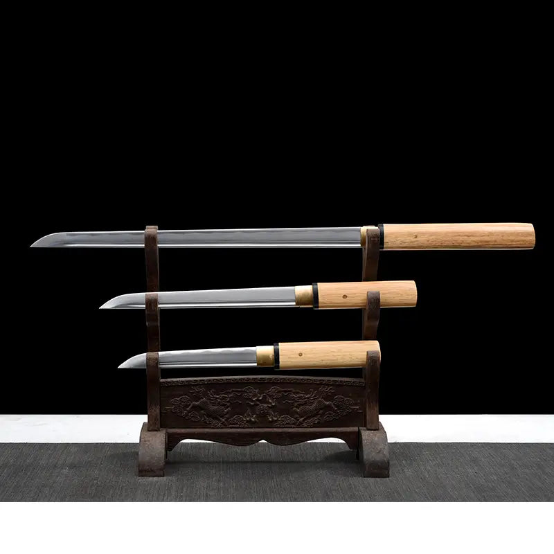 Set San-Ken : trois tantō japonais shirasaya aux lames dégainées et polies miroir, présentés sur un support en bois sculpté à trois niveaux ; poignées et fourreaux en bois clair avec bagues noires, habaki en laiton visibles, fond noir studio et tapis gris — set artisanal en acier carbone 1045.