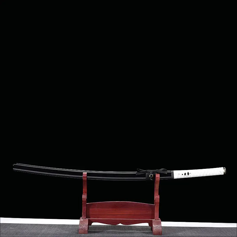 Katana japonais artisanal Shinobi no Kiba, Le Croc du Shinobi, présenté sur un support en bois rouge foncé, sur fond noir. Katana rangé dans son saya noir laqué avec sageo noir noué, tsuka en cuir blanc avec tressage ito noir, présentation horizontale mettant en valeur le profil discret et l’esthétique ninja.
