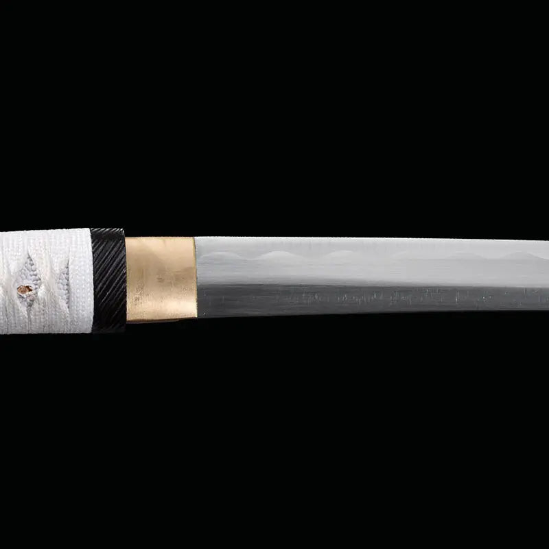 Détail de la lame du katana japonais artisanal Shinobi no Kiba, Le Croc du Shinobi, sur fond noir. Jonction entre la tsuka et la lame avec habaki doré visible, acier carbone 1045 à finition mate, lame sans bo-hi avec ligne de trempe discrète, montage sans tsuba au style ninja épuré.
