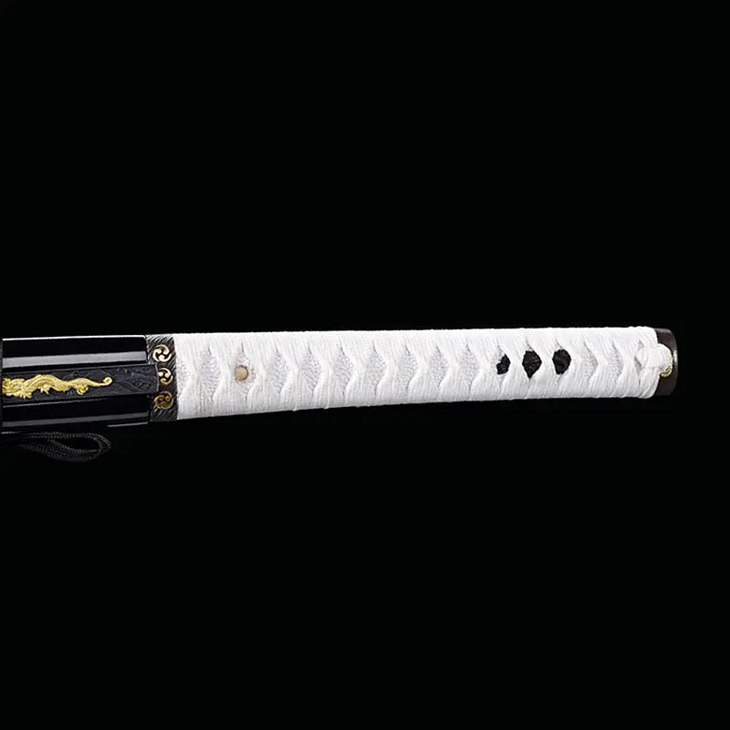 Détail de la poignée du katana japonais artisanal Shinobi no Kiba, Le Croc du Shinobi, sur fond noir. Tsuka en bois recouverte de cuir blanc avec tressage ito noir, mekugi visibles, kashira sombre, saya noir laqué partiellement visible avec décoration dorée du kozuka intégré.
