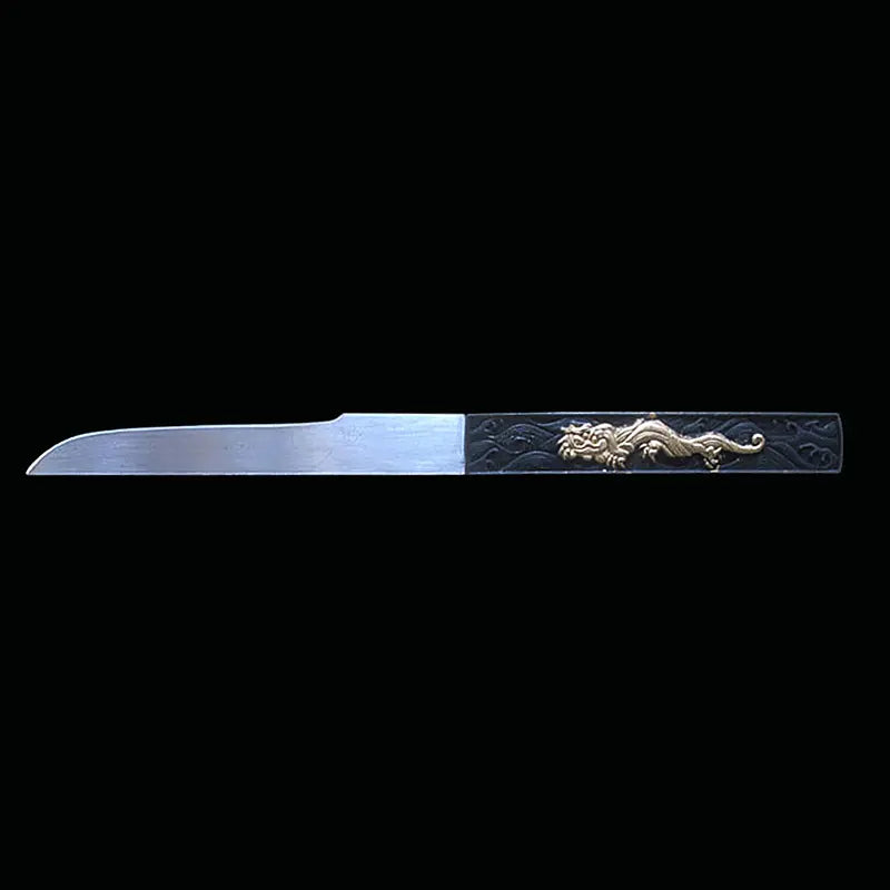 Kozuka du katana japonais artisanal Shinobi no Kiba, Le Croc du Shinobi, présenté sur fond noir. Petit couteau auxiliaire en acier inoxydable partiellement sorti de son logement, lame droite visible, manche noir avec décoration dorée en relief, intégré dans le saya noir laqué.
