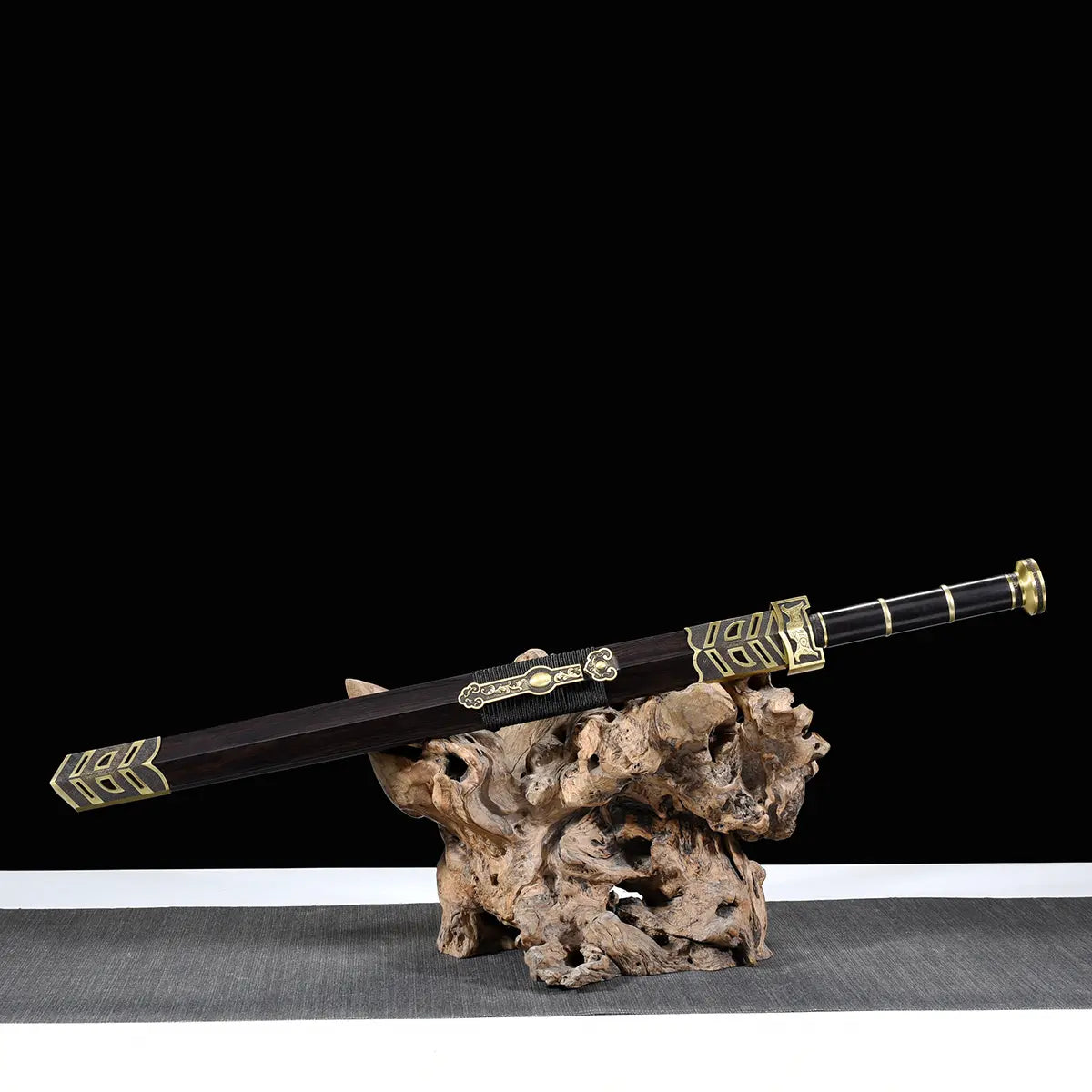 Épée chinoise Jian SHU SHENG présentée dans son fourreau noir style ébène sur un support en bois naturel, avec poignée noire, garnitures en laiton doré et ornements décoratifs visibles, posée sur une surface grise devant un fond noir.