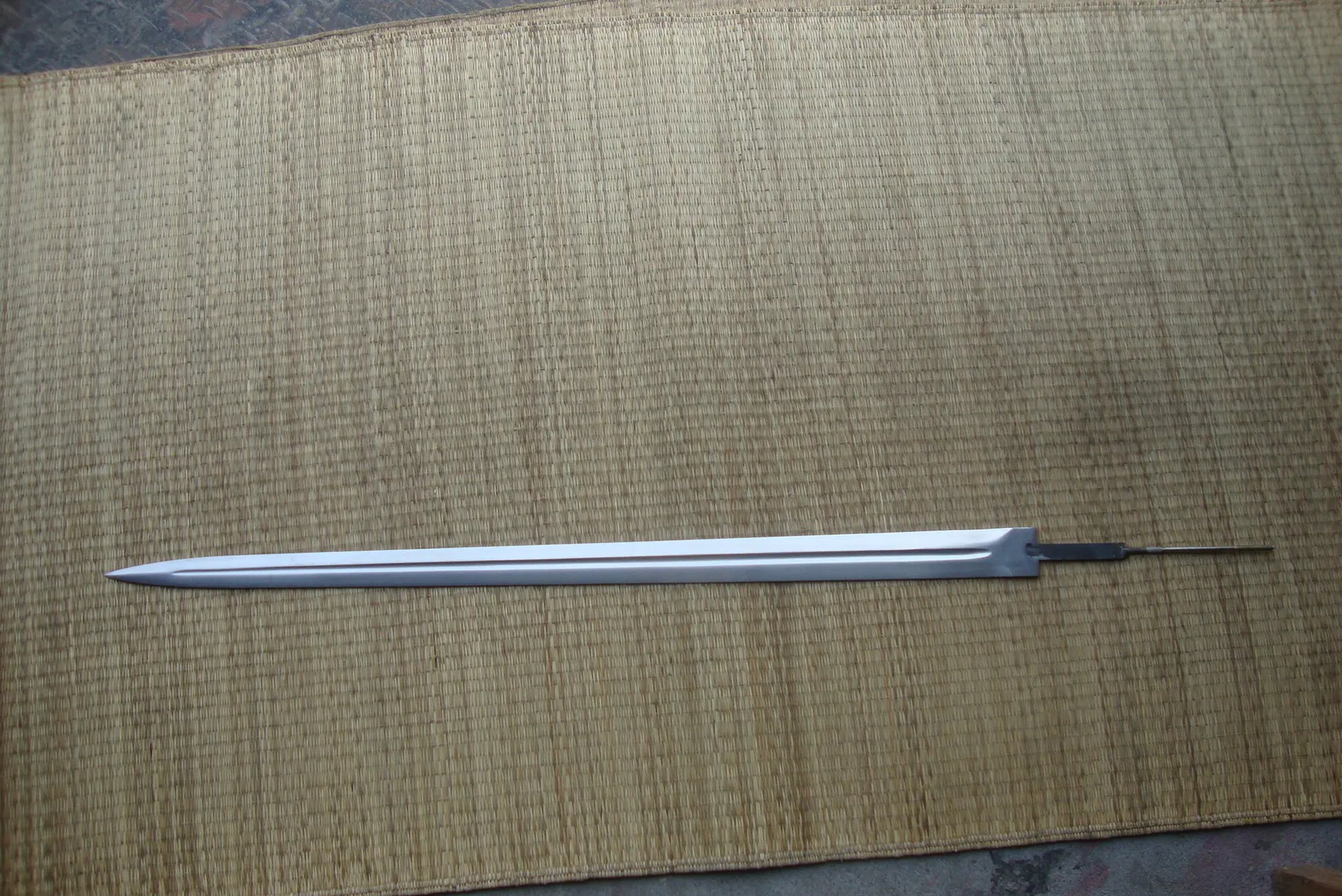 Lame de l’épée Strider inspirée d’Aragorn dans Le Seigneur des Anneaux, posée horizontalement sur un fond en natte beige tressée type tatami. Lame longue en acier inoxydable avec fuller central visible sur toute la longueur, pointe effilée et finition polie. La garde et la poignée ne sont pas montées, laissant apparaître la soie en queue de rat à l’arrière. Aucun fourreau présent sur la photo.
