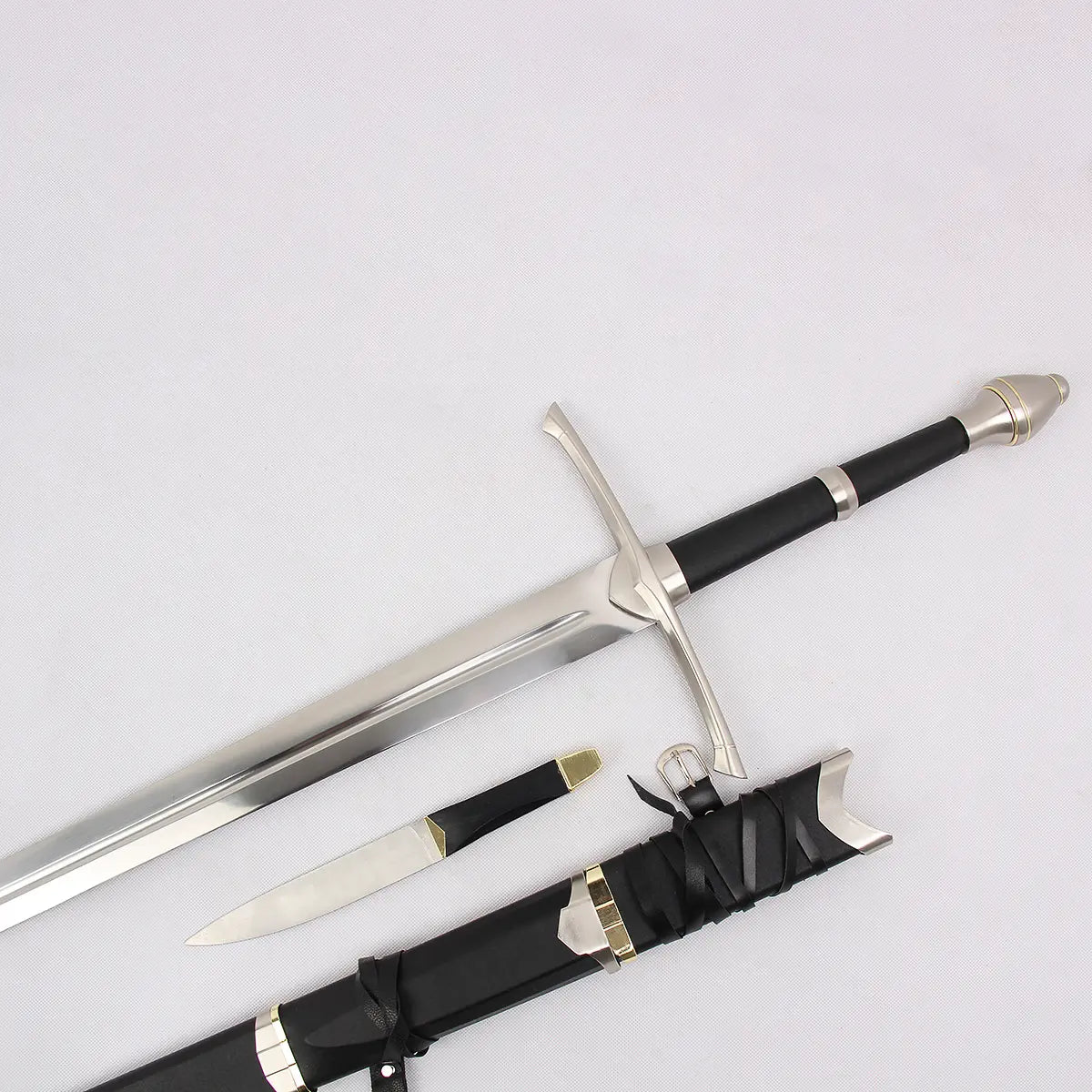 Vue rapprochée de l’épée Strider inspirée d’Aragorn dans Le Seigneur des Anneaux, photographiée sur un fond blanc texturé. La lame en acier poli avec fuller central est partiellement sortie du fourreau noir gainé de cuir. La garde en croix métallique, la poignée noire et le pommeau argenté sont visibles. Une dague assortie est placée à côté du fourreau avec des sangles en cuir et une boucle métallique apparentes.
