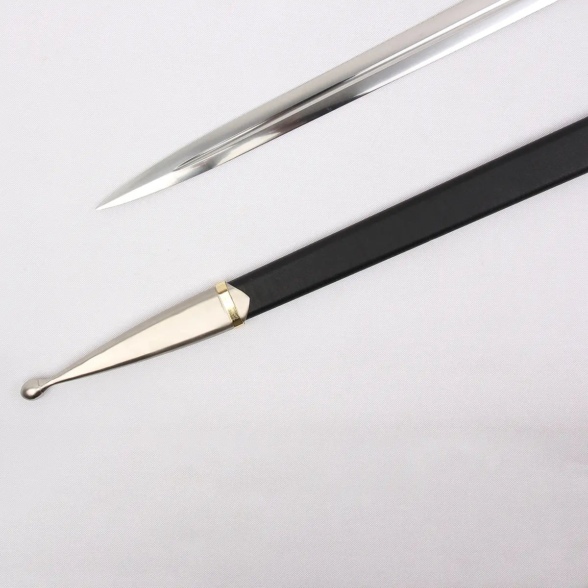 Détail de la pointe de la lame de l’épée Strider inspirée d’Aragorn dans Le Seigneur des Anneaux, photographiée sur un fond blanc texturé. La lame en acier poli présente une pointe effilée avec un fuller central visible. Le fourreau noir gainé de cuir apparaît en parallèle avec un embout métallique argenté et un annea
