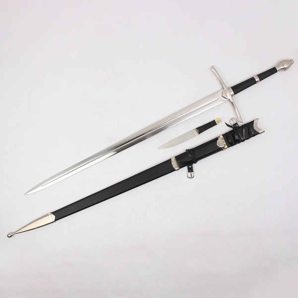 Vue complète de l’épée Strider inspirée d’Aragorn dans Le Seigneur des Anneaux, présentée en diagonale sur un fond blanc neutre. La lame en acier présente un fuller central et une finition polie. Le fourreau noir gainé de cuir est posé sous l’épée avec garnitures métalliques argentées et dorées, sangles en cuir et boucle de ceinture visibles. Une petite dague assortie est placée entre l’épée et le fourreau.
