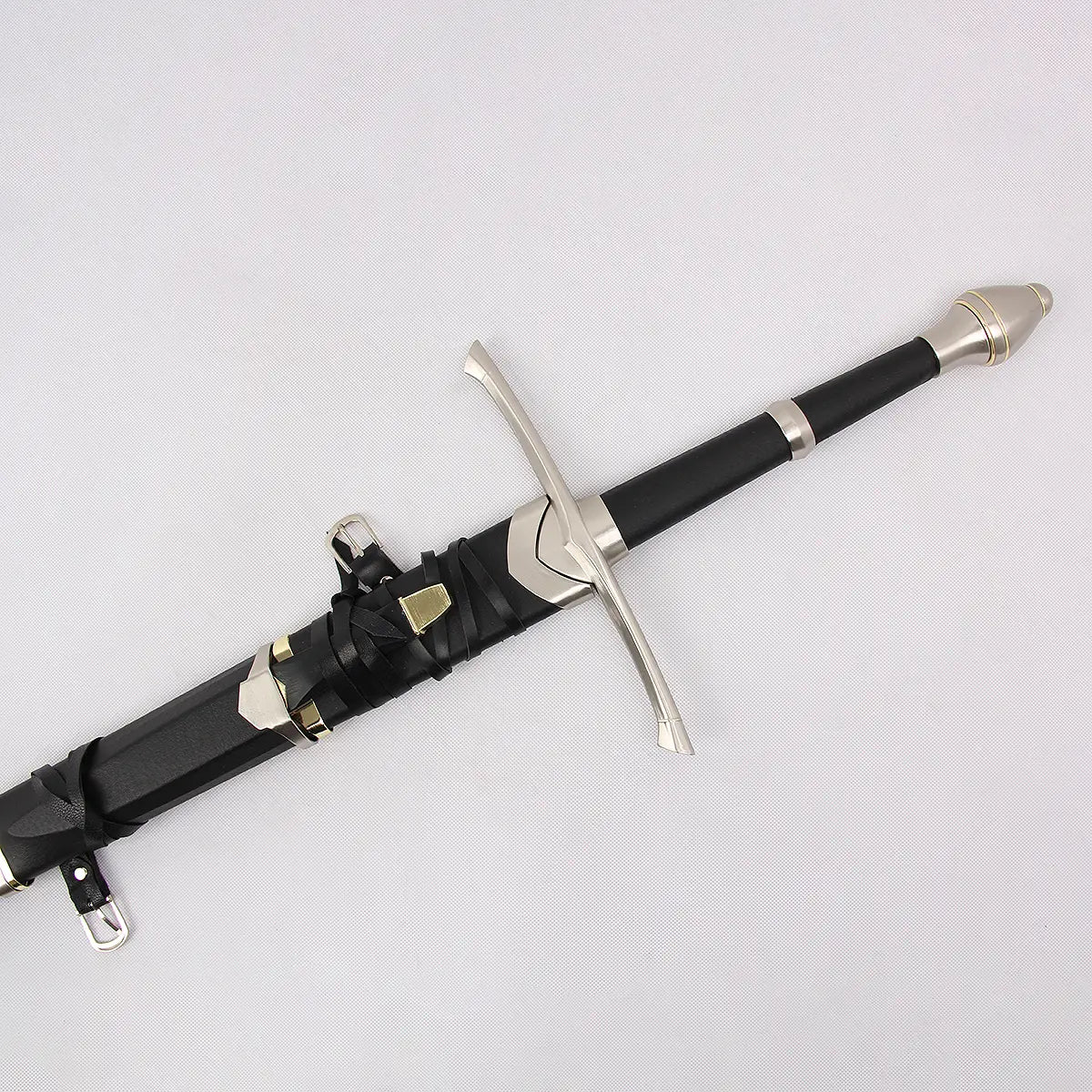 Détail de la poignée et du pommeau de l’épée Strider inspirée d’Aragorn dans Le Seigneur des Anneaux, photographiée sur un fond blanc texturé. La poignée est gainée de cuir noir avec une finition mate et entourée de bagues métalliques argentées. Le pommeau en métal présente une forme évasée avec des anneaux décoratifs dorés. La garde en croix et le fourreau noir avec sangles en cuir sont partiellement visibles.
