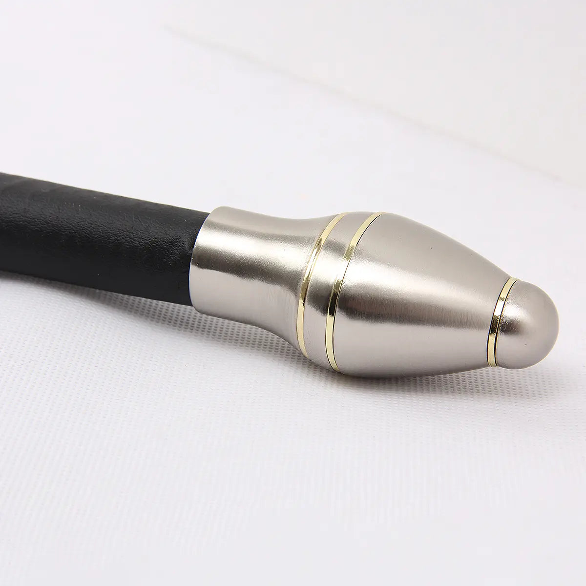 Gros plan sur le pommeau métallique de l’épée Strider inspirée d’Aragorn dans Le Seigneur des Anneaux, photographié sur un fond blanc texturé. Le pommeau argenté présente une forme arrondie et légèrement évasée avec deux anneaux décoratifs dorés. La poignée est gainée de cuir noir à texture mate, sans fourreau visible dans ce cadrage.
