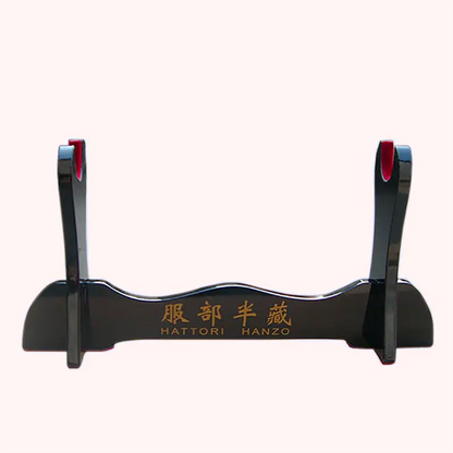 Support katana en bois laqué noir, inscriptions dorées « HATTORI HANZO » avec kanji, coussinets rouges sur les bras, photographié de face en packshot sur fond uni rose pâle, sans sabre — présentoir pour sabre japonais / support katana décoratif.