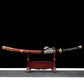 Full view of the Japanese tachi katana Taiyo Taisho displayed horizontally on a traditional red wooden sword stand, photographed against a deep black background with a neutral gray surface below, showing the red lacquered saya with gold fittings, braided sageo cord, black tsuka with white ray skin panels and gold menuki, studio lighting emphasizing balance, curvature, and premium craftsmanship of the complete katana presentation
