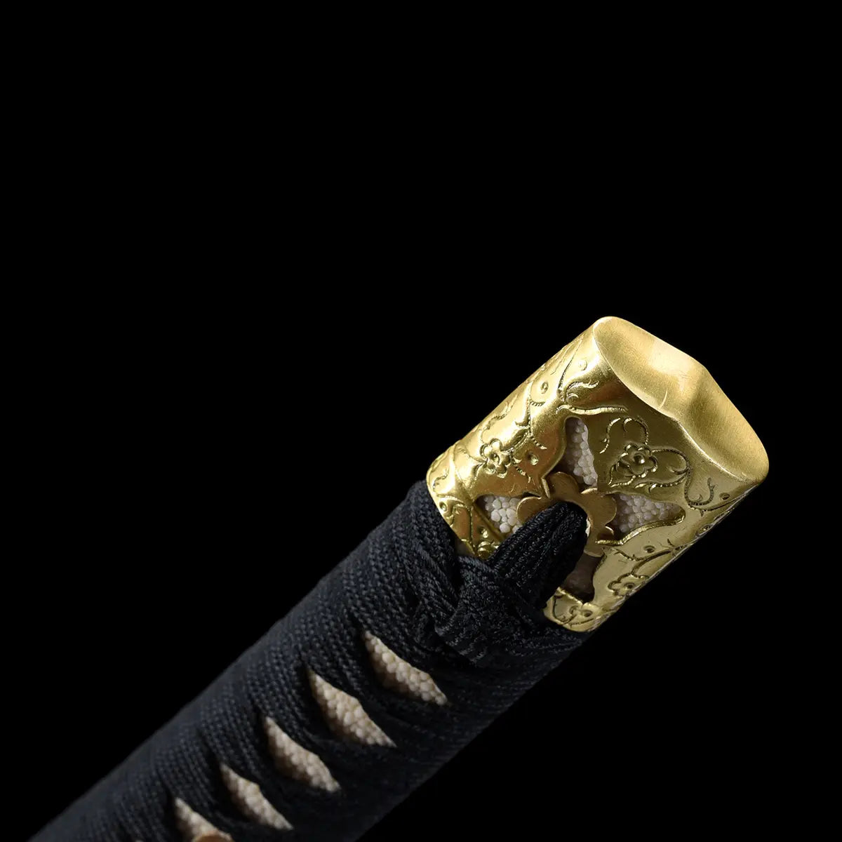 Close-up detail of the Japanese tachi katana Taiyo Taisho photographed on a deep black background, focusing on the handle end with a gold-colored ornate kashira engraved with traditional motifs, black tsuka tightly wrapped in black ito over white ray skin panels, visible texture of the wrap and metal engraving, studio lighting highlighting the craftsmanship and refined finish of the handle pommel
