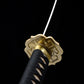 Close-up view of the Japanese tachi katana Taiyo Taisho photographed on a deep black background, focusing on the gold-colored ornate tsuba and brass habaki, with the blade extending upward out of frame, black tsuka tightly wrapped in black ito over white ray skin panels, fine engraved details visible on the guard and collar, studio lighting emphasizing the metal finish and traditional Japanese craftsmanship
