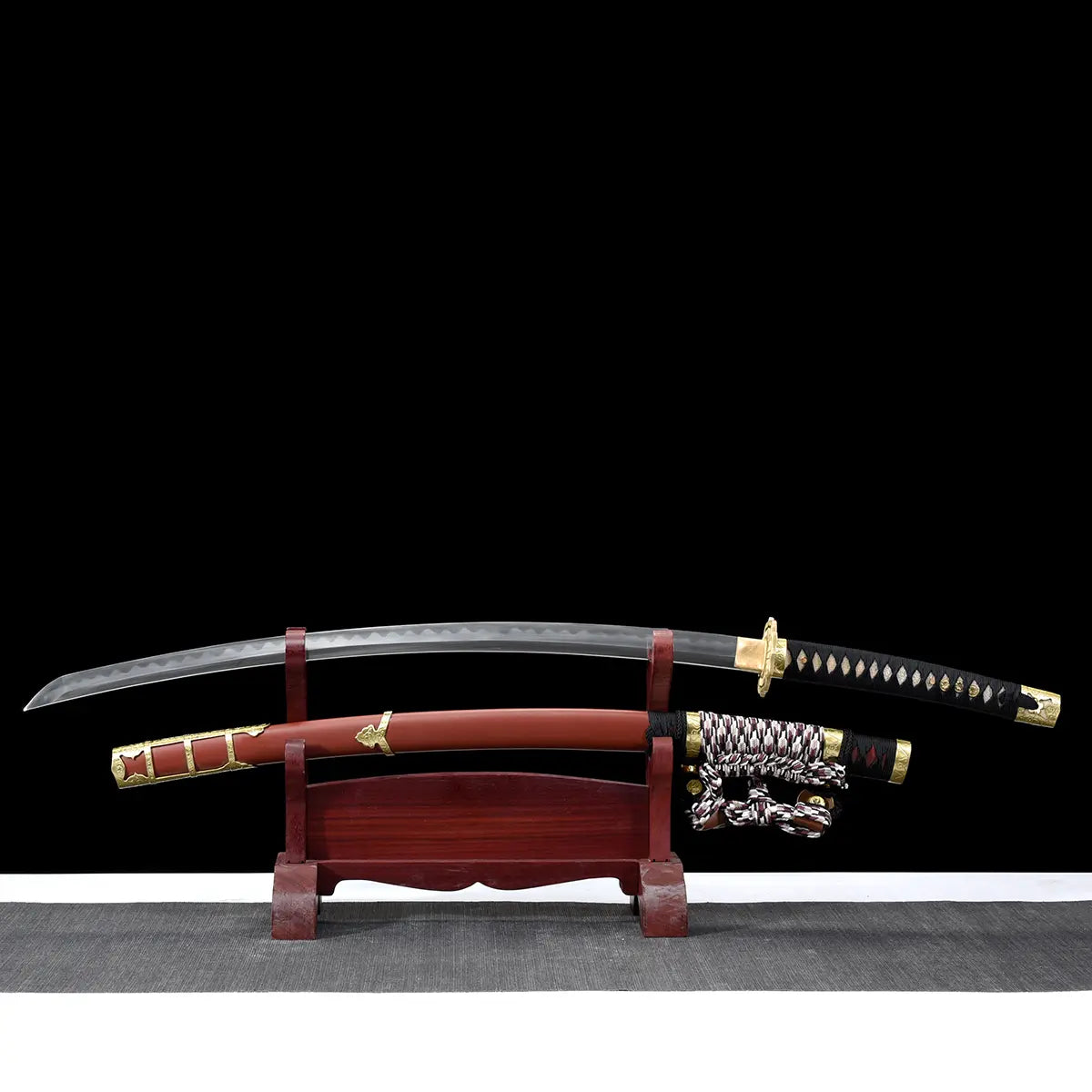 Unsheathed Japanese tachi katana Taiyo Taisho displayed on a traditional red wooden sword stand, showing the curved steel blade with visible hamon resting above the red lacquered saya with gold fittings, braided sageo cord positioned near the handle, black tsuka with white ray skin panels and gold menuki, photographed in studio on a deep black background with a gray surface, highlighting blade curvature, balance, and premium Japanese craftsmanship
