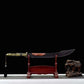 Sabre chinois Dao TIAN DAO présenté dans son fourreau noir courbe sur un support en bois rouge, avec poignée dorée rétro-futuriste, garde métallique, pompon suspendu, fond noir et figurine d’éléphant décorative à droite.