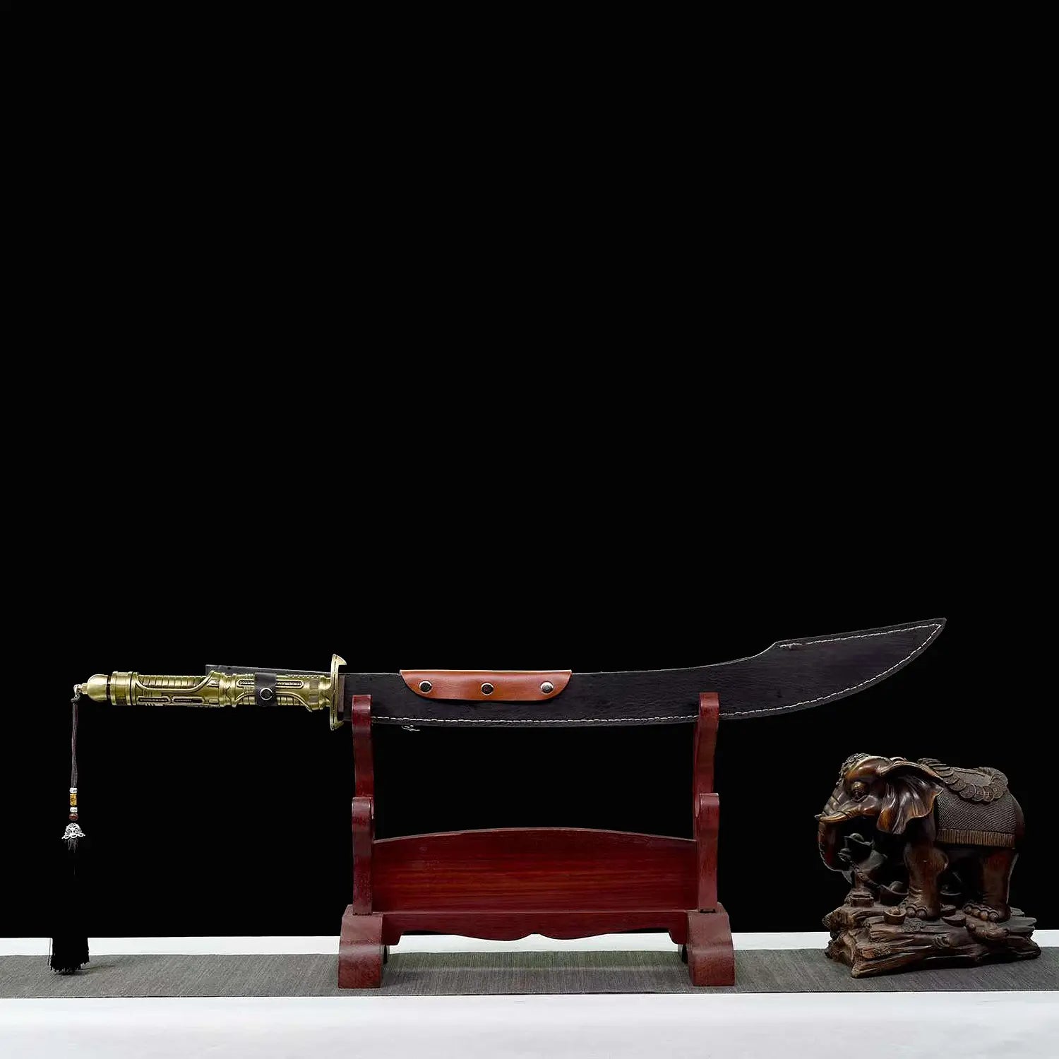Sabre chinois Dao TIAN DAO présenté dans son fourreau noir courbe sur un support en bois rouge, avec poignée dorée rétro-futuriste, garde métallique, pompon suspendu, fond noir et figurine d’éléphant décorative à droite.