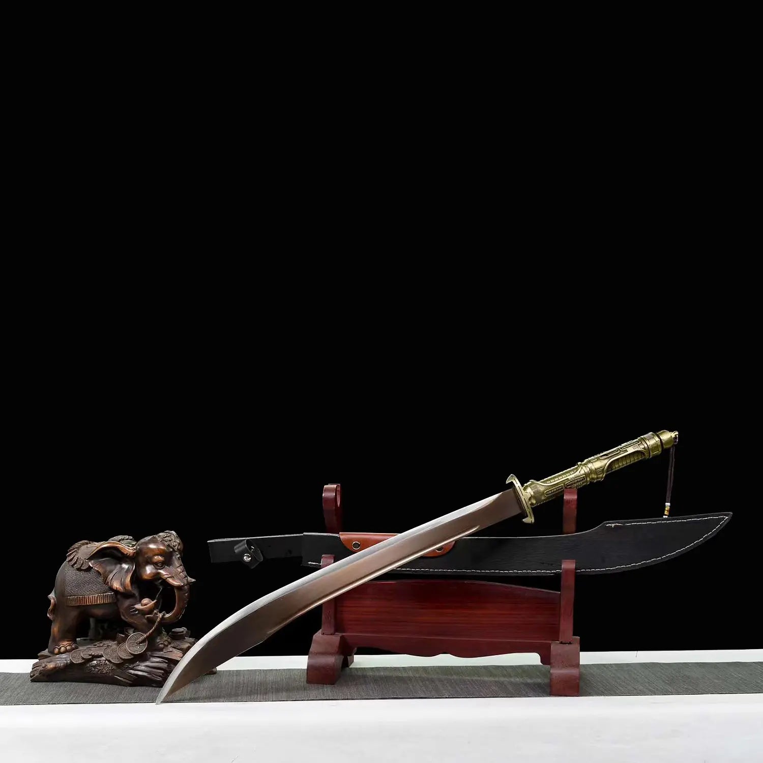 Sabre chinois Dao TIAN DAO présenté en diagonale avec lame courbe sortie, poignée dorée rétro-futuriste, fourreau noir à coutures blanches, support en bois rouge et figurine d’éléphant décorative sur fond noir.