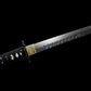 Gros plan du wakizashi Hoshizora : habaki en laiton gravé devant tsuba ronde noire, début de lame acier T10 trempe à l’argile montrant la texture martelée tsuchime et le hamon ondulé ; poignée (tsuka) en ito noir sur samegawa blanc, menuki visibles ; prise de vue sur fond noir — sabre artisanal Blade Bushido.