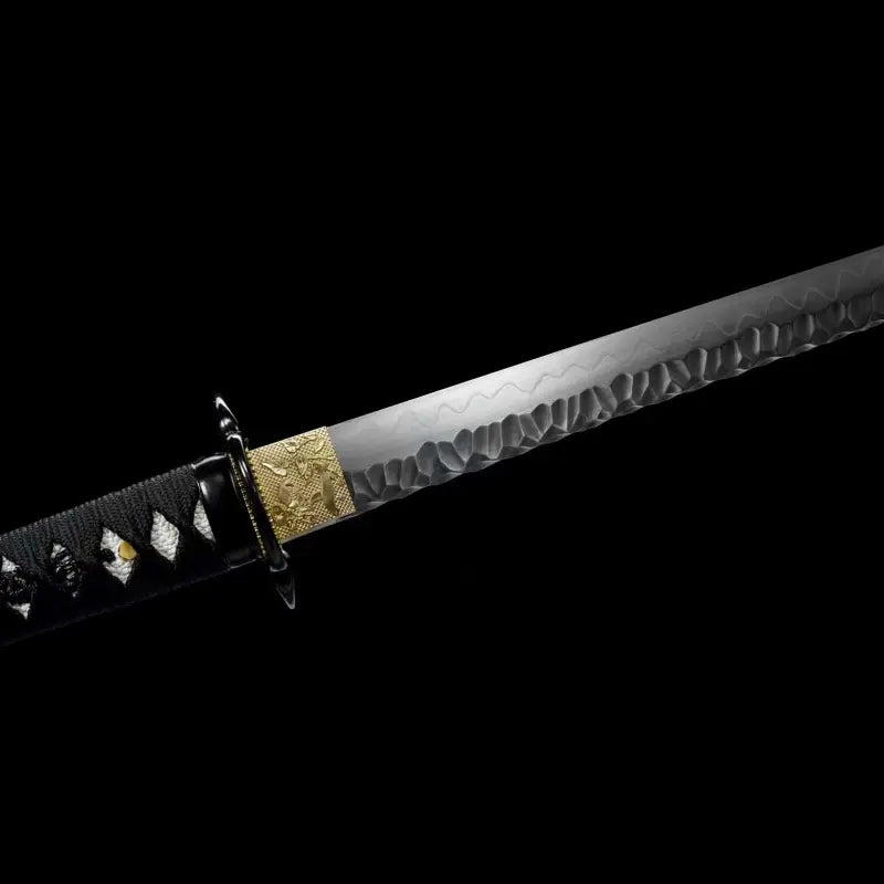 Gros plan du wakizashi Hoshizora : habaki en laiton gravé devant tsuba ronde noire, début de lame acier T10 trempe à l’argile montrant la texture martelée tsuchime et le hamon ondulé ; poignée (tsuka) en ito noir sur samegawa blanc, menuki visibles ; prise de vue sur fond noir — sabre artisanal Blade Bushido.