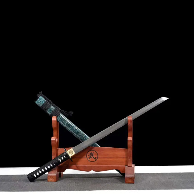 Wakizashi japonais artisanal Aonami avec lame T10 sans bohi posée en diagonale sur un support en bois (kanji gravé), hamon visible, habaki en laiton, tsuka noire à losanges de same blanche ; saya nashiji bleu-vert et sageo noir à l’arrière-plan, fond noir et tapis gris.