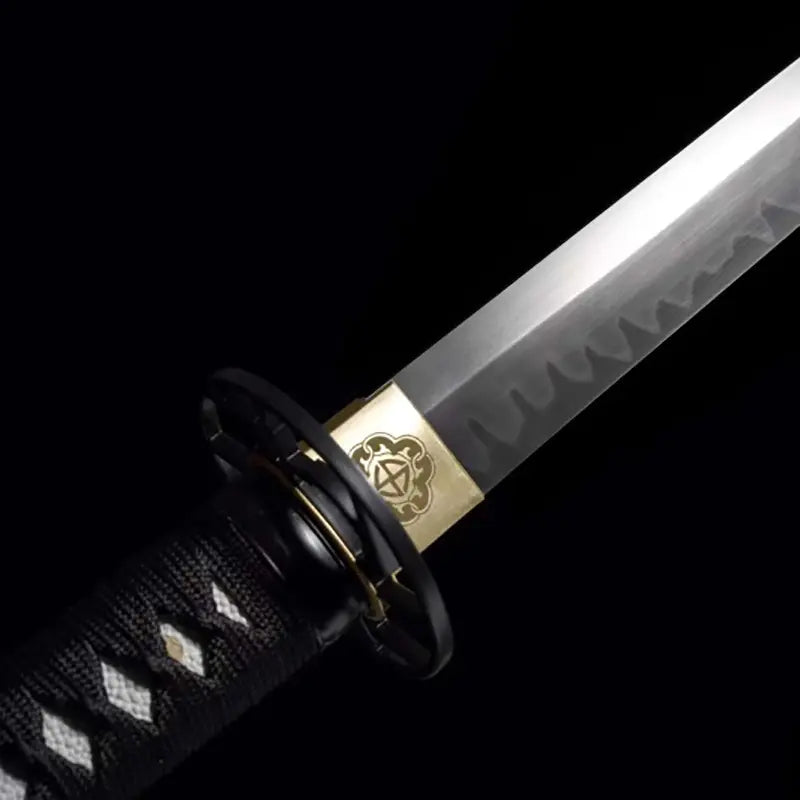 Gros plan du wakizashi japonais artisanal Aonami : habaki en laiton gravé, tsuba ronde noire, hamon ondé bien visible sur la lame T10 sans bohi ; tsuka en ito noir à losanges de same blanche, photographié sur fond noir pour un rendu haut contraste.