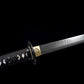 Gros plan du wakizashi japonais artisanal Aonami : tsuka en ito noir à losanges de same blanche, tsuba ronde noire, habaki en laiton gravé et lame T10 sans bohi montrant un hamon ondé ; prise de vue latérale sur fond noir.