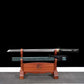 Wakizashi japonais artisanal Aonami en acier T10, lame dégainée sans bohi avec hamon visible, posé sur un support en bois (saya nashiji bleu-vert au niveau inférieur), tsuka noire à losanges de same blanche, tsuba ronde noire et habaki laiton, sur fond noir avec tapis gris