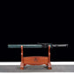 Wakizashi japonais artisanal Aonami en fourreau : saya nashiji bleu-vert avec sageo noir, tsuka noire à losanges de same blanche, posé horizontalement sur un support en bois (kanji au centre), fond noir et surface tapis gris — acier T10, trempe à l’argile, hamon réel.