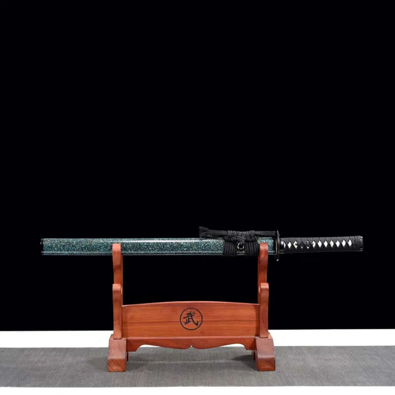 Wakizashi japonais artisanal Aonami en fourreau : saya nashiji bleu-vert avec sageo noir, tsuka noire à losanges de same blanche, posé horizontalement sur un support en bois (kanji au centre), fond noir et surface tapis gris — acier T10, trempe à l’argile, hamon réel.