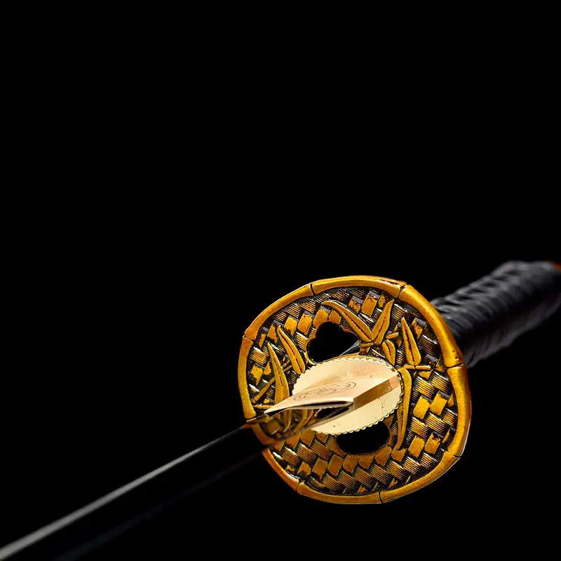 Gros plan de la tsuba dorée motif bambou du wakizashi japonais “Kurochiku”, habaki en laiton au premier plan, début de lame noire satinée, tsuka noire en arrière-plan flou, sur fond noir profond.