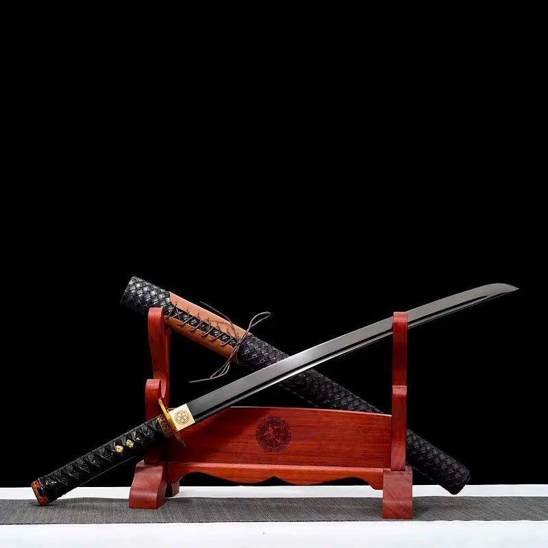 Wakizashi japonais “Kurochiku” présenté en diagonale sur un support en bois rouge : lame noire satinée avec bohi bien visible, tsuba dorée motif bambou, tsuka noire à menuki dorés, saya tressé noir en arrière-plan ; scène posée sur tapis gris avec fond noir uniforme.