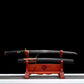 Wakizashi japonais “Kurochiku” dégainé sur présentoir en bois rouge : lame noire satinée avec bohi au-dessus du saya tressé noir (laçage marron), tsuka noire à menuki dorés, posé sur un tapis gris avec arrière-plan noir uniforme.