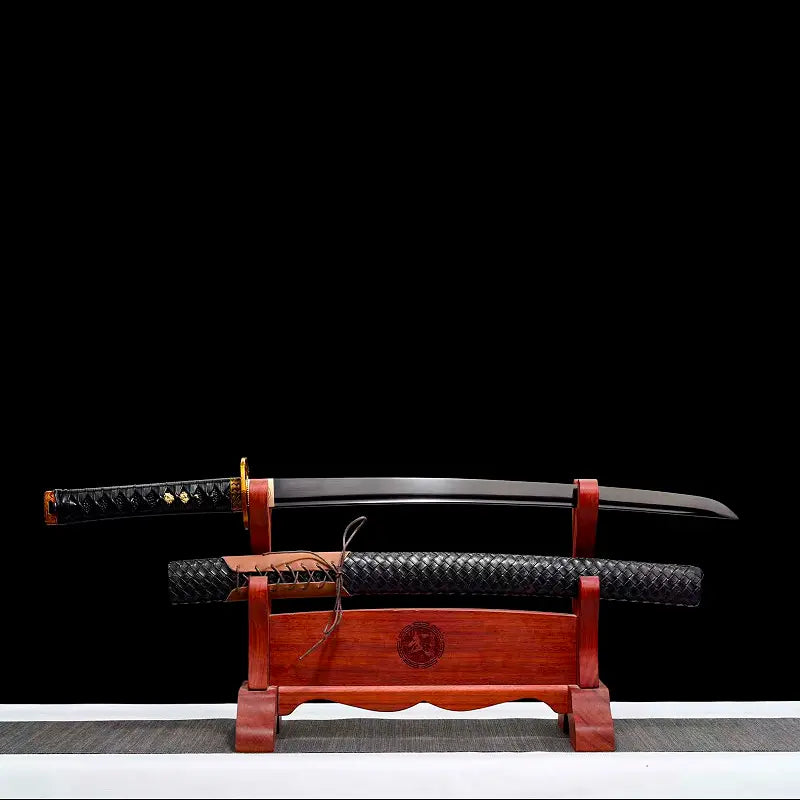 Wakizashi japonais “Kurochiku” dégainé sur présentoir en bois rouge : lame noire satinée avec bohi au-dessus du saya tressé noir (laçage marron), tsuka noire à menuki dorés, posé sur un tapis gris avec arrière-plan noir uniforme.
