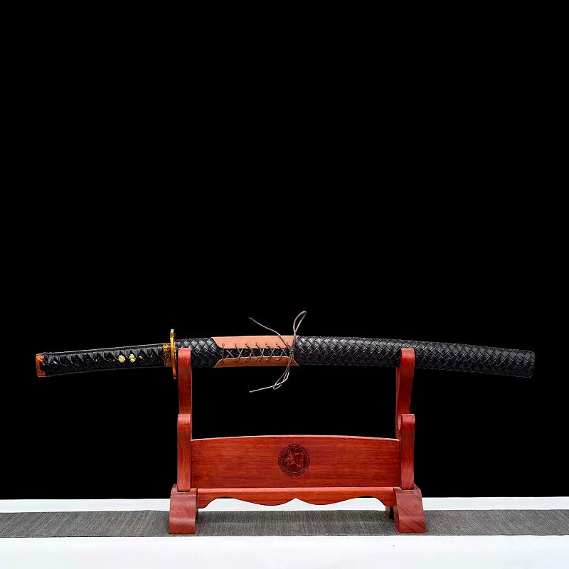 Wakizashi japonais “Kurochiku” au repos sur un support en bois rouge, saya noir à tressage façon cuir avec laçage marron au centre, tsuka noire visible côté gauche ; scène posée sur un tapis gris, fond noir uniforme pour un rendu épuré et premium.