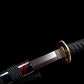 Gros plan lors de l’extraction : wakizashi japonais Renge no Reicha, habaki gravé de feuilles et tsuba en laiton motif lotus, tsuka tressé noir, saya noire laquée à liserés pourpres, lame acier plié damassé mise en lumière sur fond noir — sabre court artisanal, Blade Bushido.