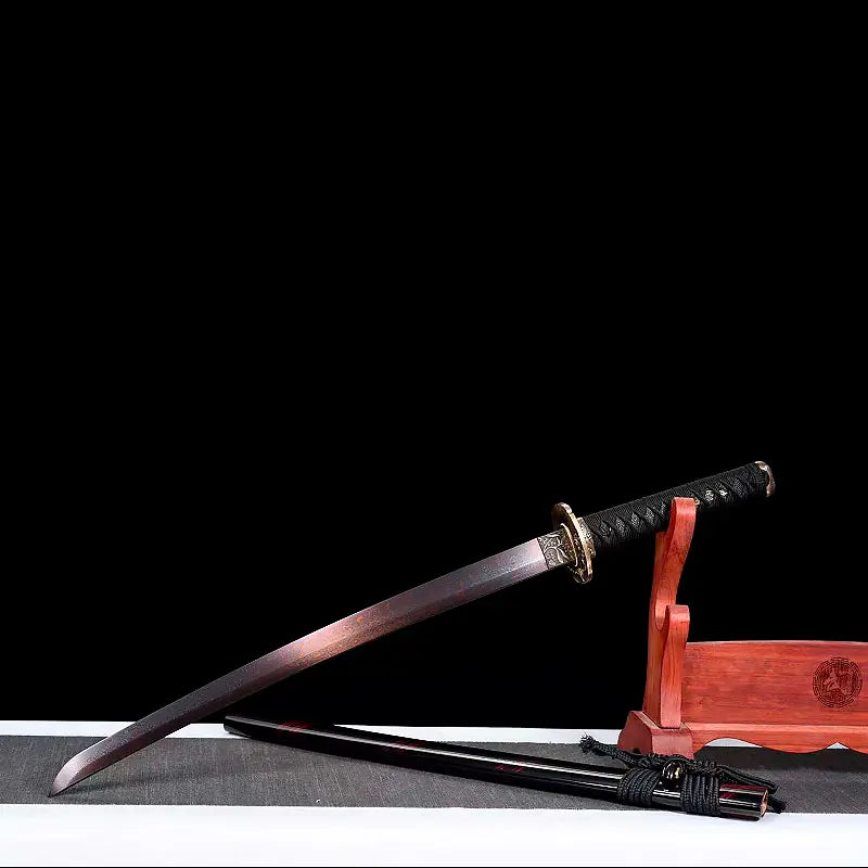 Wakizashi japonais Renge no Reicha posé en biais sur toile grise, lame acier plié damassé à patine sombre, tsuba laiton motif lotus et habaki gravé, saya noire laquée aux liserés pourpres et sageo noir à l’avant, support en bois verni à droite, fond noir studio — sabre court artisanal Blade Bushido.