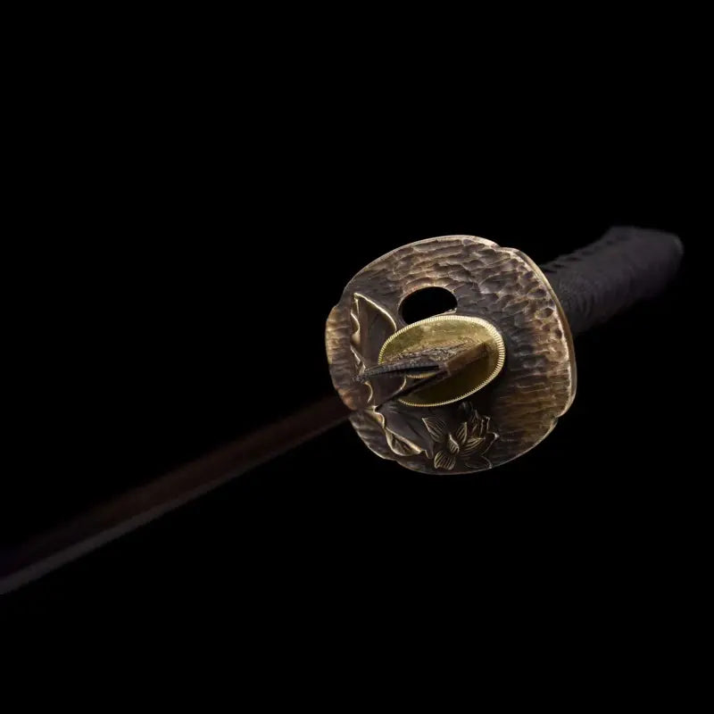Gros plan de la tsuba en laiton motif lotus du wakizashi japonais Renge no Reicha, tsuka tressé noir, début de lame sombre en perspective, éclairage doux sur fond noir studio — sabre court artisanal acier plié damassé, Blade Bushido.