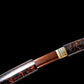 Gros plan en diagonale de la jonction habaki en laiton entre le saya et la tsuka d’un wakizashi shirasaya artisanal Kuro Yōgan ; lame acier plié motif damas rouge-noir légèrement dégainée, laque urushi noir-vermillon brillante sur les deux parties, fond noir studio, détail de montage premium.