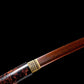 Gros plan du habaki en laiton sculpté et de la lame acier plié motif damas rouge-noir avec bohi visible, sortant du saya laqué urushi noir-vermillon ; cadrage diagonal en studio fond noir, finition brillante, style shirasaya artisanal.