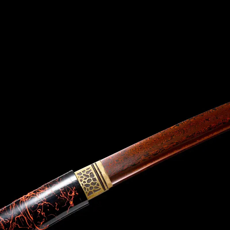 Gros plan du habaki en laiton sculpté et de la lame acier plié motif damas rouge-noir avec bohi visible, sortant du saya laqué urushi noir-vermillon ; cadrage diagonal en studio fond noir, finition brillante, style shirasaya artisanal.